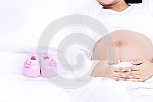 Pregnant woman lying on white bed with pink baby shoes at home.Close up abdomen people with child shoes in bedroom
