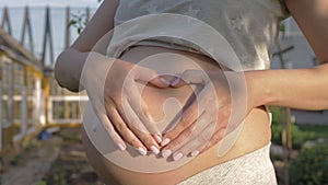 Pregnant woman makes a heart on her stomach with her hands in light of sunset