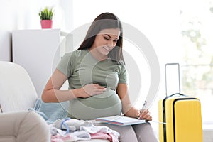 Pregnant woman making list while packing suitcase for maternity hospital