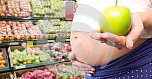 Pregnant woman mid section holding apple in blurry vegetable aisle