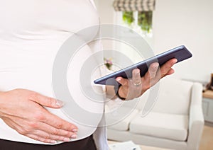 Pregnant woman mid section holding tablet against blurry sitting room