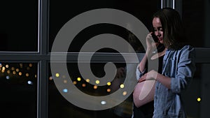 A pregnant woman in the night talking on the mobile phone hands touching the stomach standing at the window