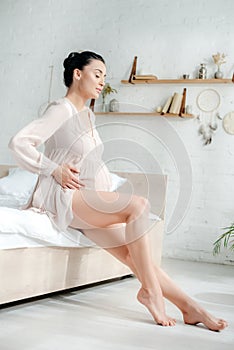 Pregnant woman in nightie touching tummy while sitting on bed