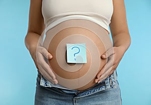 Pregnant woman with note on belly against light blue background, closeup. Choosing baby name