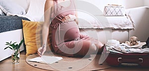 Pregnant woman packing bag for maternity hospital, making notes, checking list in diary. Expectant mother with suitcase of baby