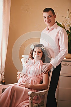 Pregnant woman in a pink dress sitting in a chair behind a smiling man standing. expecting a baby