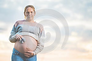 Pregnant woman posing outdoors holding little baby shoes on her belly.