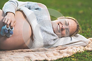 Pregnant woman posing outdoors holding little baby shoes on her belly.