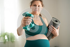 A pregnant woman prepare for exercising with dumbbells. Young girl holding a yoga mat in hand.