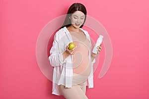 Pregnant woman with pretty stomach holding green apple and bottle of milk, expectant mother looking down, posing isolated over