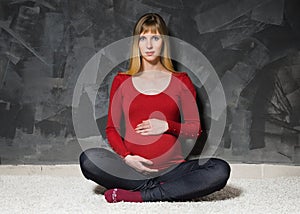 Pregnant woman in red jacket sitting near grey wall