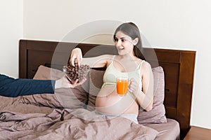 Pregnant woman relaxing in bed is eating chocolate cereal balls and drinking juice. Husband is feeding his wife. Dry breakfast