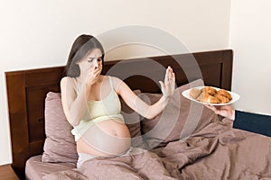 Pregnant woman relaxing in bed makes stop gesture to croissants. Expecting mother refuses to eat pastry. Diet during pregnancy