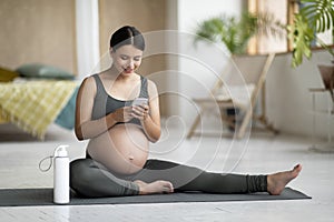 Pregnant woman relaxing with smartphone on yoga mat after training at home