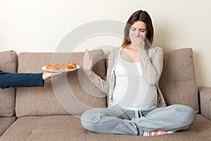 Pregnant woman relaxing on the sofa makes stop gesture to croissants. Expecting mother refuses to eat pastry. Diet during