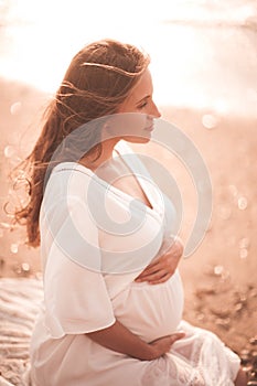 Pregnant woman showing heart shape with fingers wearing white dress over sea background. Motherhood. Maternity.