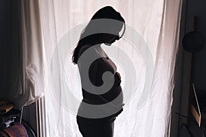 Pregnant woman silhouette standing against the window. intimate portrait expecting woman touching her big belly. concept of