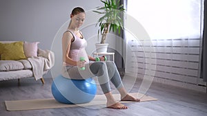 Pregnant woman sit on fit ball, working out with dumbbells in home living room. Maternity