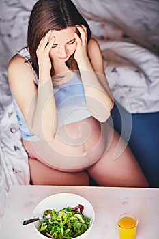 A pregnant woman sits on the bed and holds her head. Depression during pregnancy is a very common phenomenon. Headaches and lack o