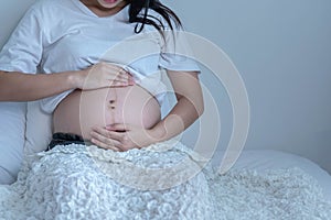 A pregnant woman sits on the bed. She uses both hands to touch and hold the fetus.