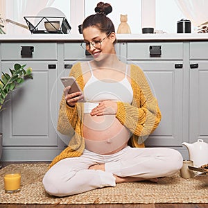 A pregnant woman sits on the kitchen floor and looks at her smartphone