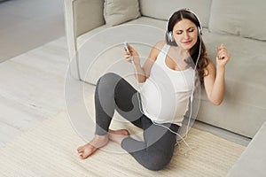 A pregnant woman sits on a light floor at home. She wore headphones and listened to music