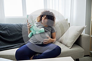A pregnant woman sits on a sofa with a child& x27;s toy rabbit for Easter in her hands smile. Lifestyle of a pregnant