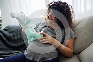 A pregnant woman sits on a sofa with a child& x27;s toy rabbit for Easter in her hands smile. Lifestyle of a pregnant