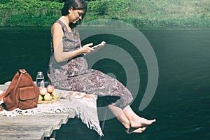 Pregnant woman sitting barefoot on wooden pier near lake and reading e-book. Outdoor picnic near water. Wellbeing and time to