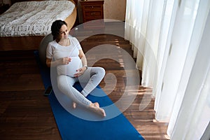 Pregnant woman sitting barefoot on yoga mat stroking her big belly, relaxing after prenatal relaxation exercises at home