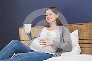 Pregnant woman sitting in bed with a cup of beverage with one hand on her belly