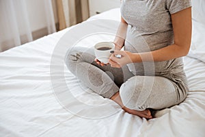 Pregnant woman sitting on bed and drinking coffee at home