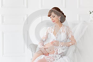 Pregnant woman sitting in a chair in a beautiful white dress boudoir .