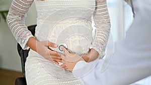 Pregnant woman sitting on chair while the doctor checking her stomach, using stethoscope