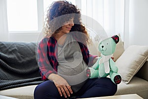 A pregnant woman is sitting on the couch with a children& x27;s toy rabbit in her hands talking to the child for Easter