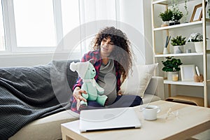 A pregnant woman is sitting on the couch with a children& x27;s toy rabbit in her hands talking to the child for Easter