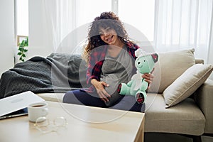 A pregnant woman is sitting on the couch with a children& x27;s toy rabbit in her hands talking to the child for Easter