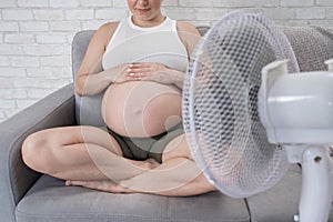 A pregnant woman is sitting on the couch, stroking her tummy and enjoying the cool air from an electric fan.