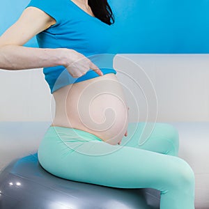 Pregnant woman sitting on fit ball, pointing on her belly