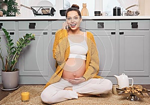 A pregnant woman is sitting on the floor of her kitchen, talking on her cell phone