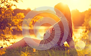 Pregnant woman sitting on green grass, looking on river on sunset, touching her belly. Healthy pregnancy