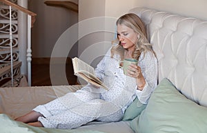Pregnant woman sitting at home in bed, reading book and drinking tea or water from cup
