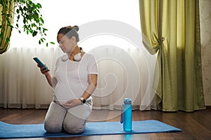 Pregnant woman sitting on mat, strokes her belly, adjusts soundtrack and mobile app with pregnancy yoga her smartphone