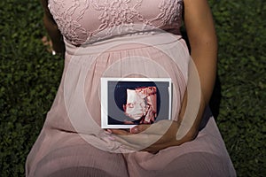 Pregnant woman sitting in the park on the grass holding 3D  image ultrasound of baby in her womb