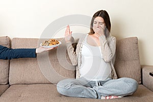 Pregnant woman sitting on the sofa refuses to eat cookies and makes no gesture. Healthy diet during pregnancy concept