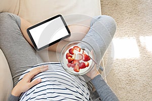 Pregnant woman sitting sofa using digital tablet while eating yogurt