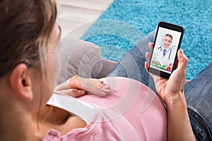 Pregnant Woman Sitting On Sofa Video Conferencing With Doctor