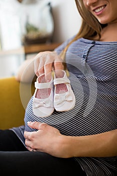 Pregnant Woman Sitting On Sofa At Home Holding Baby Shoes