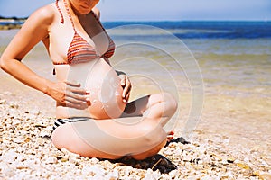 Pregnant woman skin care moisturizer cream on belly on beach at