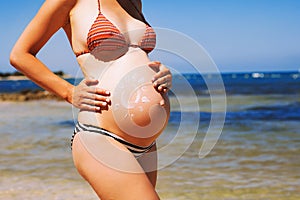 Pregnant woman skin care moisturizer cream on belly on beach at